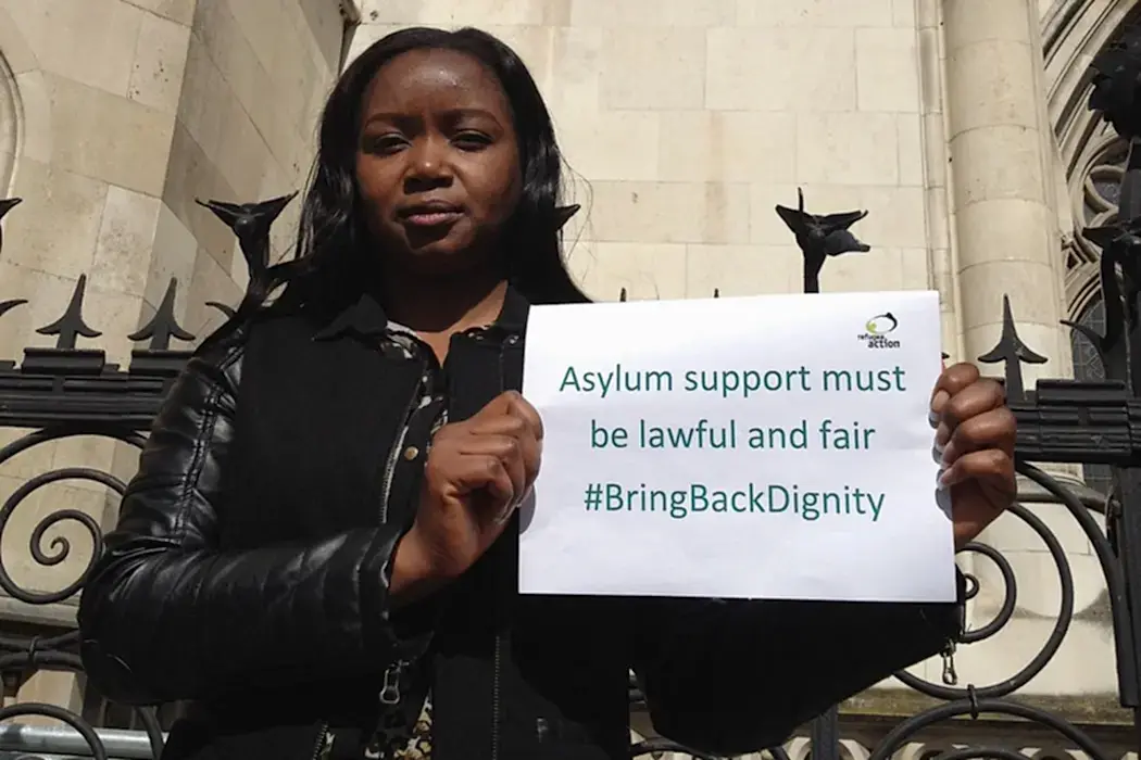 A young woman stands next to a iron railing outside a stone courthouse building, holding a paper sign which reads, "Asylum support must be lawful and fair. #BringBackDignity."