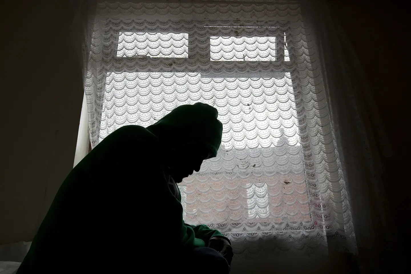 In silhouette, a young asylum seeker sits by a window in his accommodation.