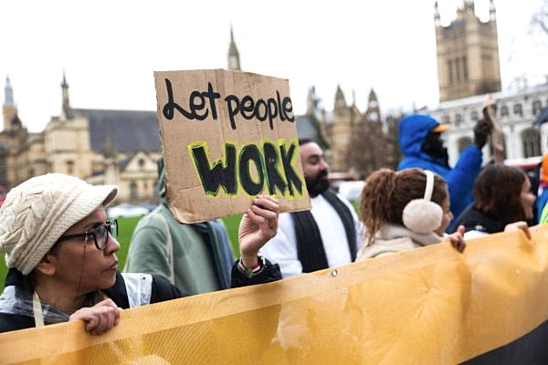A group of people stand holding a huge banner, and a sign saying 'let people work'