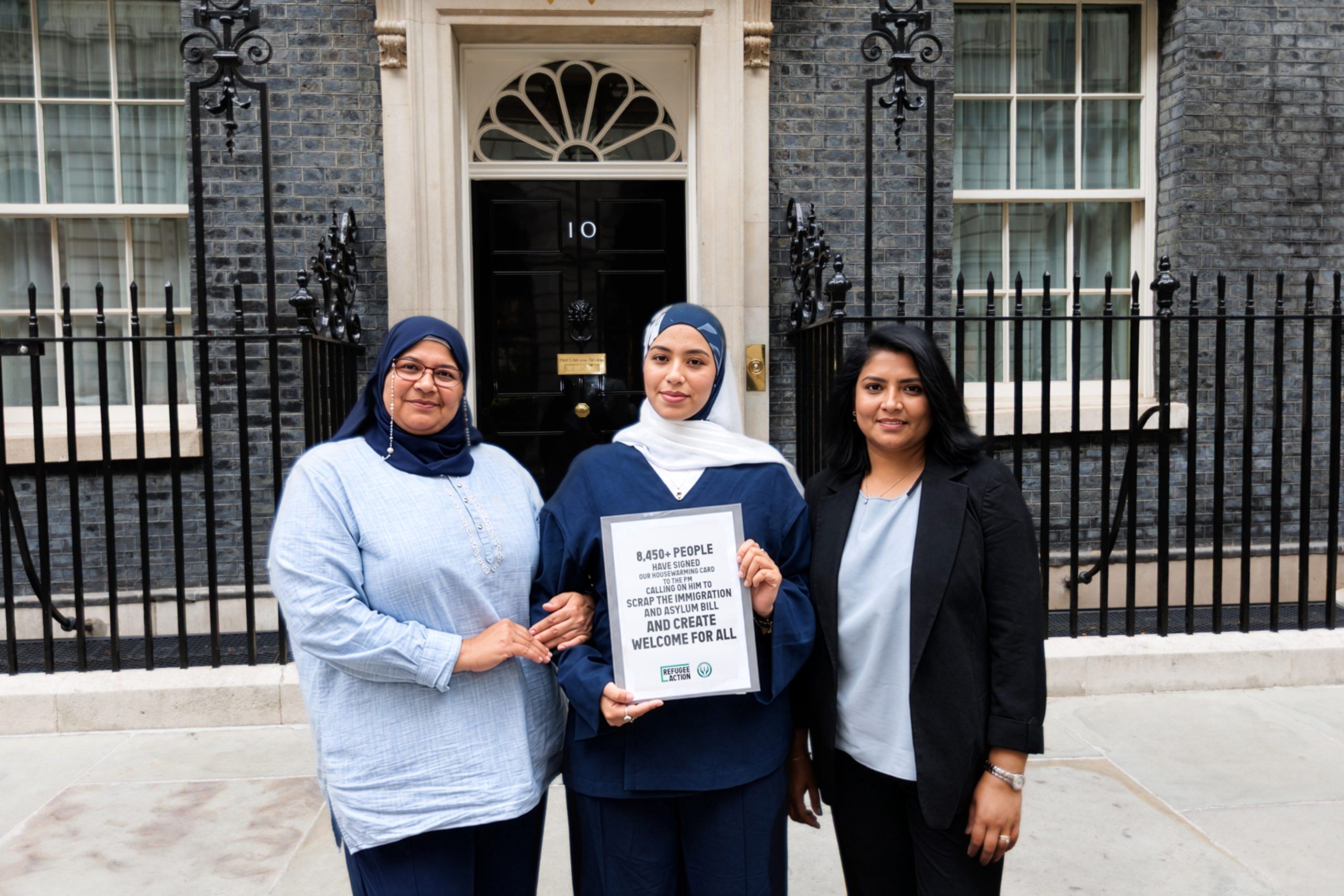 Three women are smiling powrfully at the camera, holding out the folder which says "8450 people signed our housewarming card to the PM calling on him to scrap the immigration and asylum bill and create welcome for all".