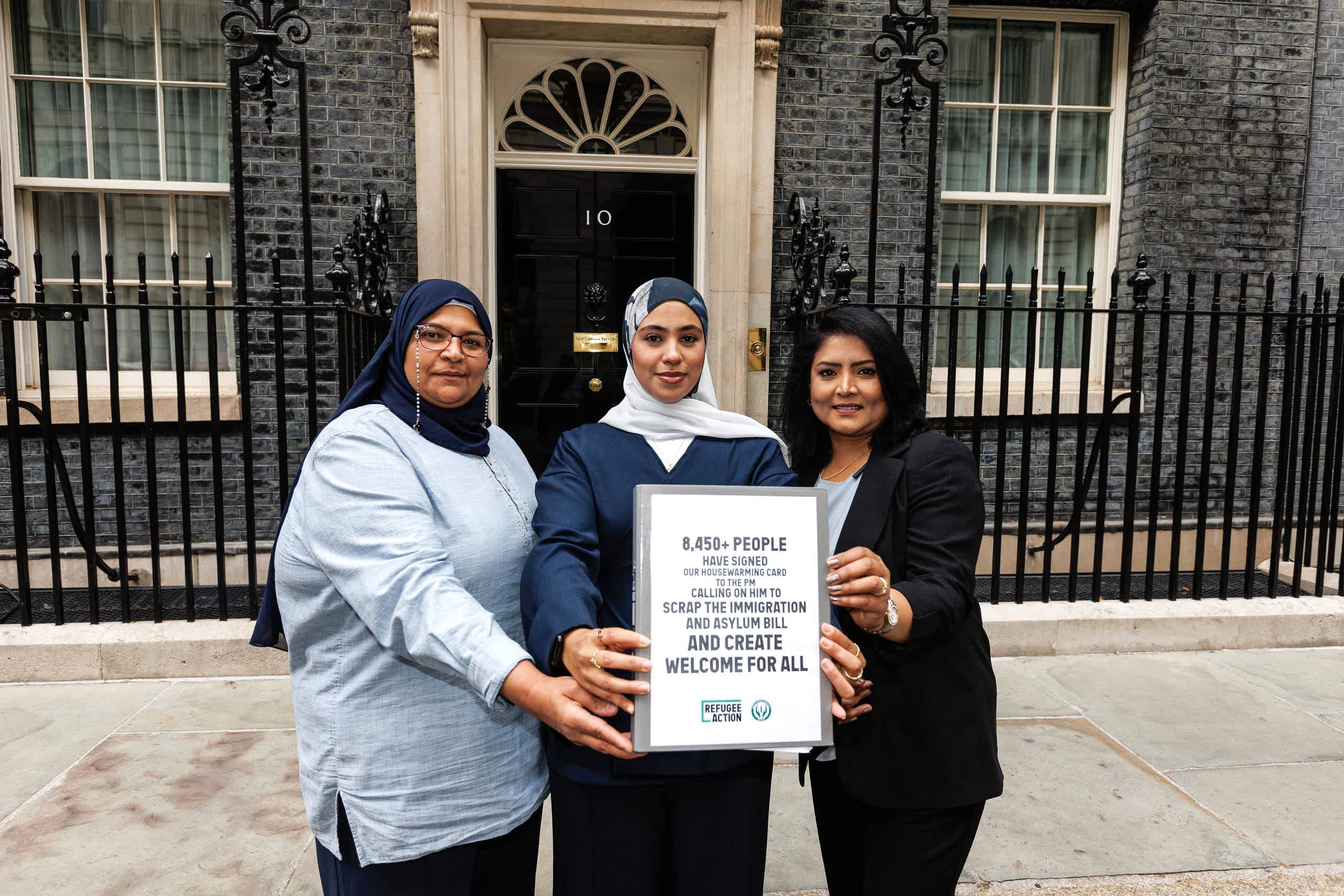 Three women are smiling powrfully at the camera, holding out the folder which says "8450 people signed our housewarming card to the PM calling on him to scrap the immigration and asylum bill and create welcome for all".