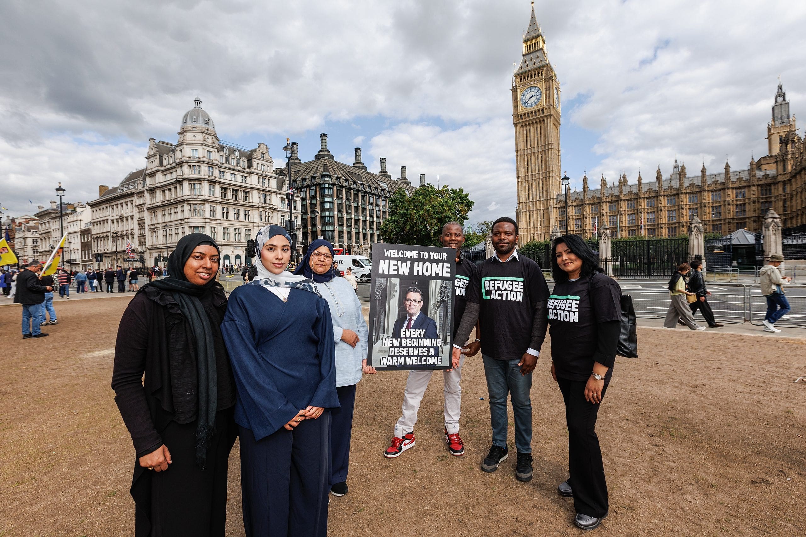 Campaigners gather on the lawn at parliament square with Big Ben behind them. They are holding a giant version of the housewarming card which says "every new beginning deserves a warm welcome"