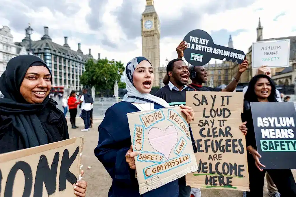 Campaigners cheering outside parliament with signs. One of the says "asylum means safety"