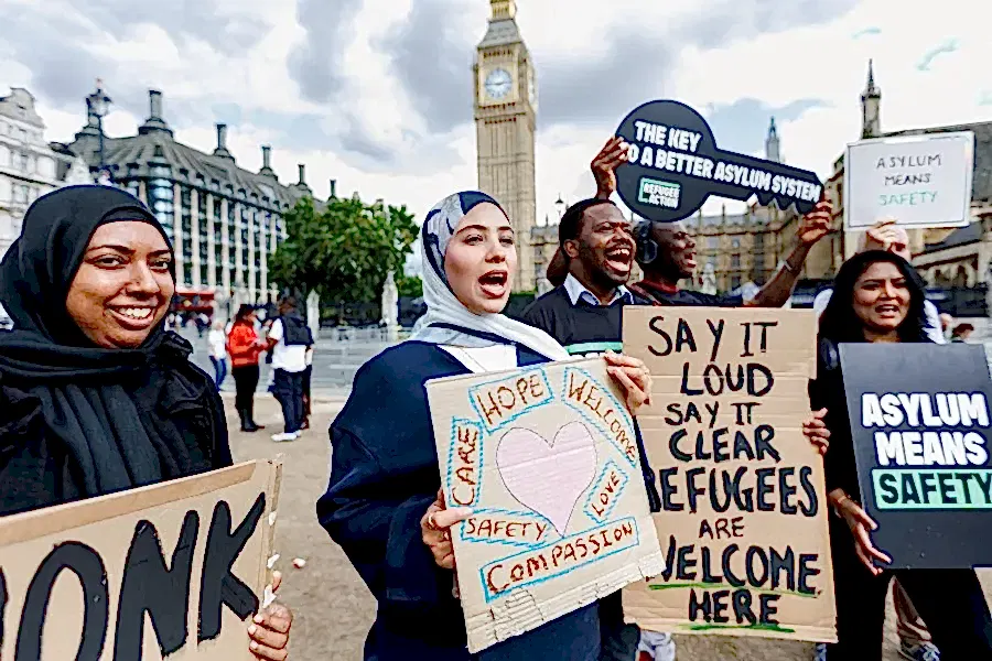 Campaigners cheering outside parliament with signs. One of the says "asylum means safety"