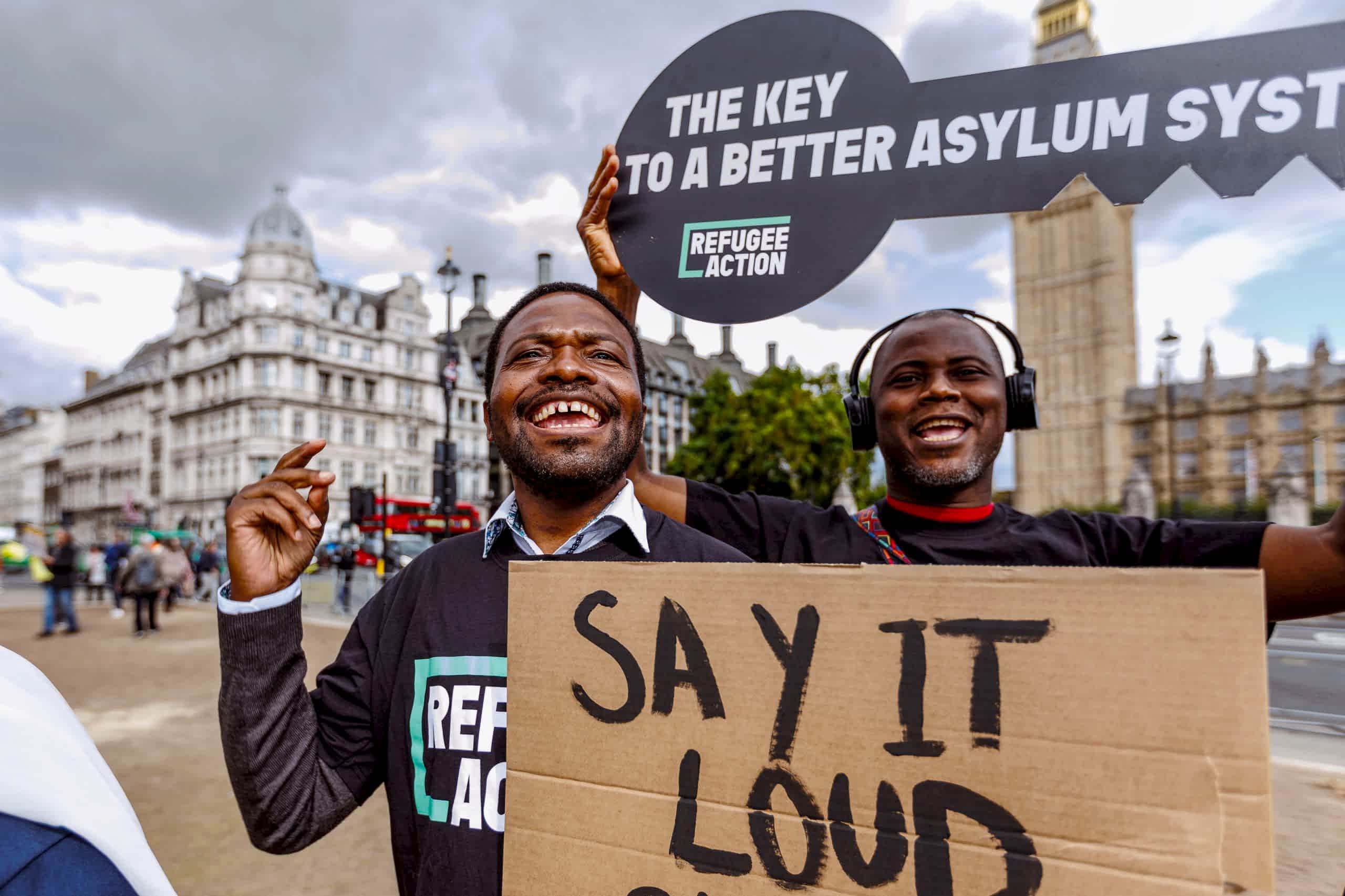 Jonathan and his fellow campaigner look elated, waving signs which say "a key to a better asylum system". Big ben is behind the,