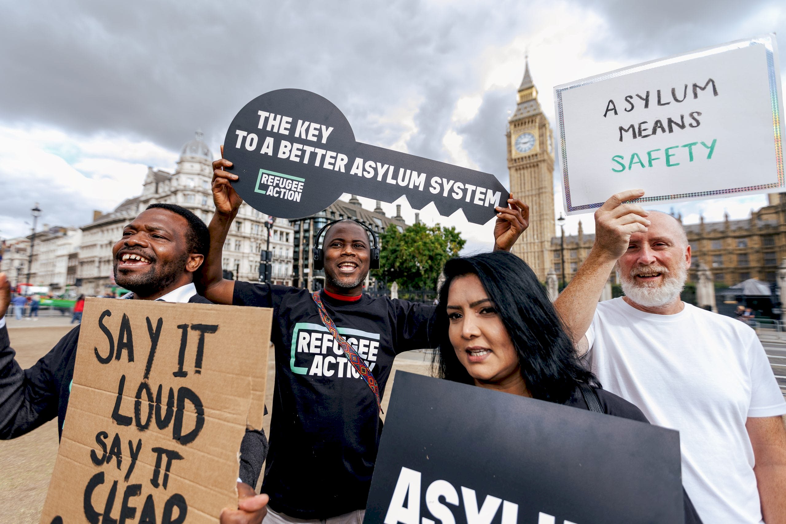 Campaigners are gathered on Parliament Square with Big Ben behind them, all holding signs. One of the signs says "say it loud, say it clear, refugees are welcome here"