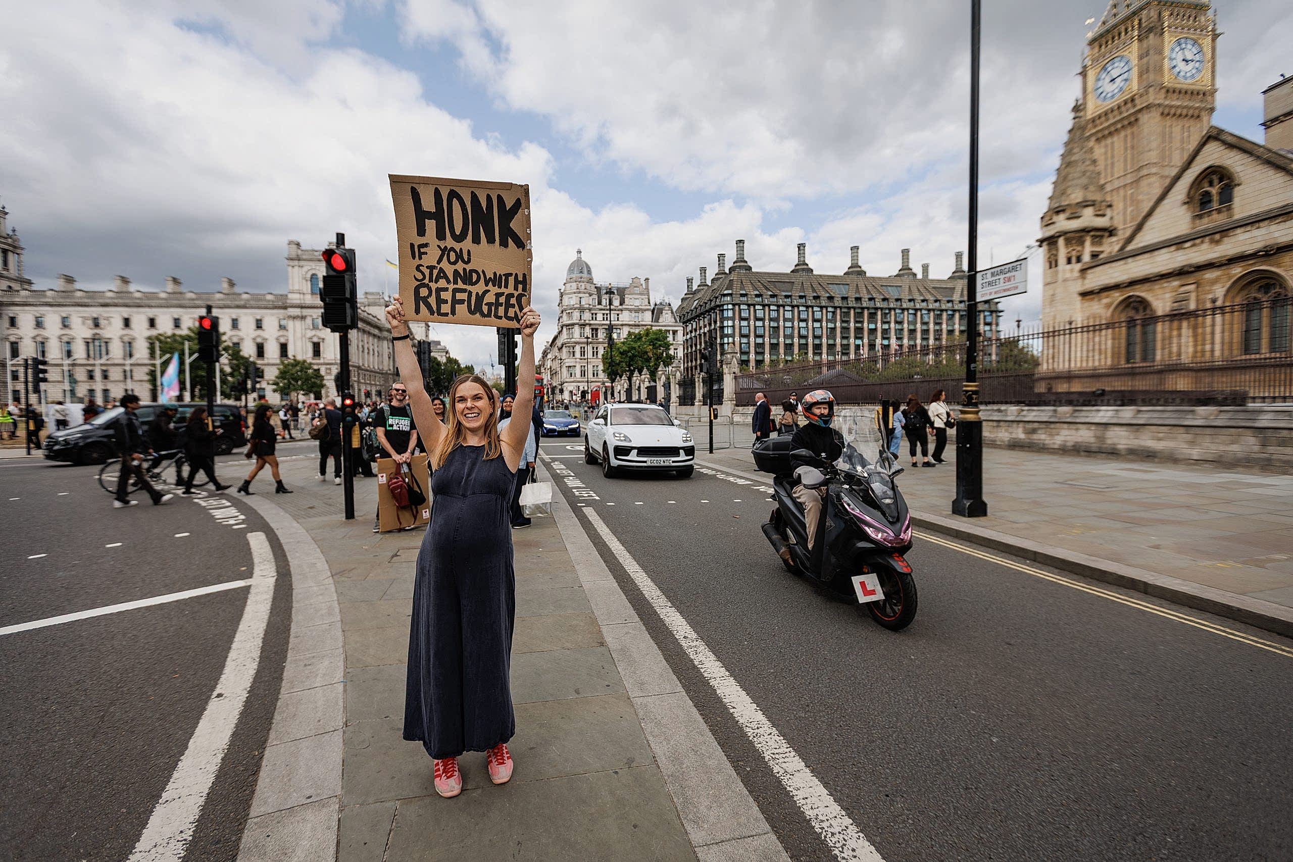 Cars drive past as Bronya holds a sign in the air which says "honk if you stand with refugees". Shes smiling down the camera, and wearing a black jumpsuit with pink shoes.