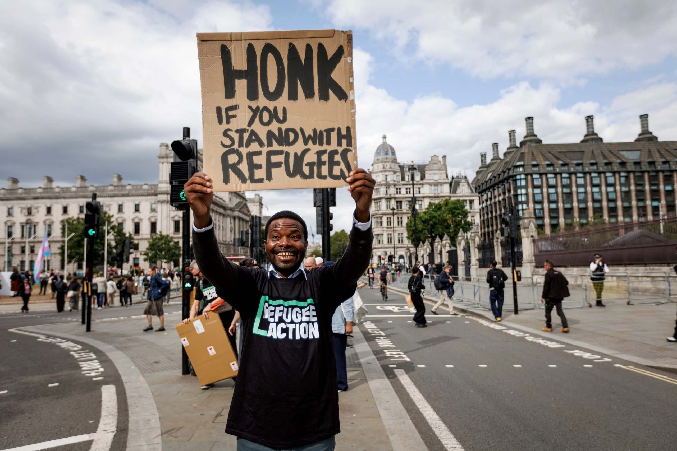 Jonathan is beaming down the camera with such a big gin. He is wearing a refugee action tshirt, and holding a sign which says "Honk if you stand with refugees".