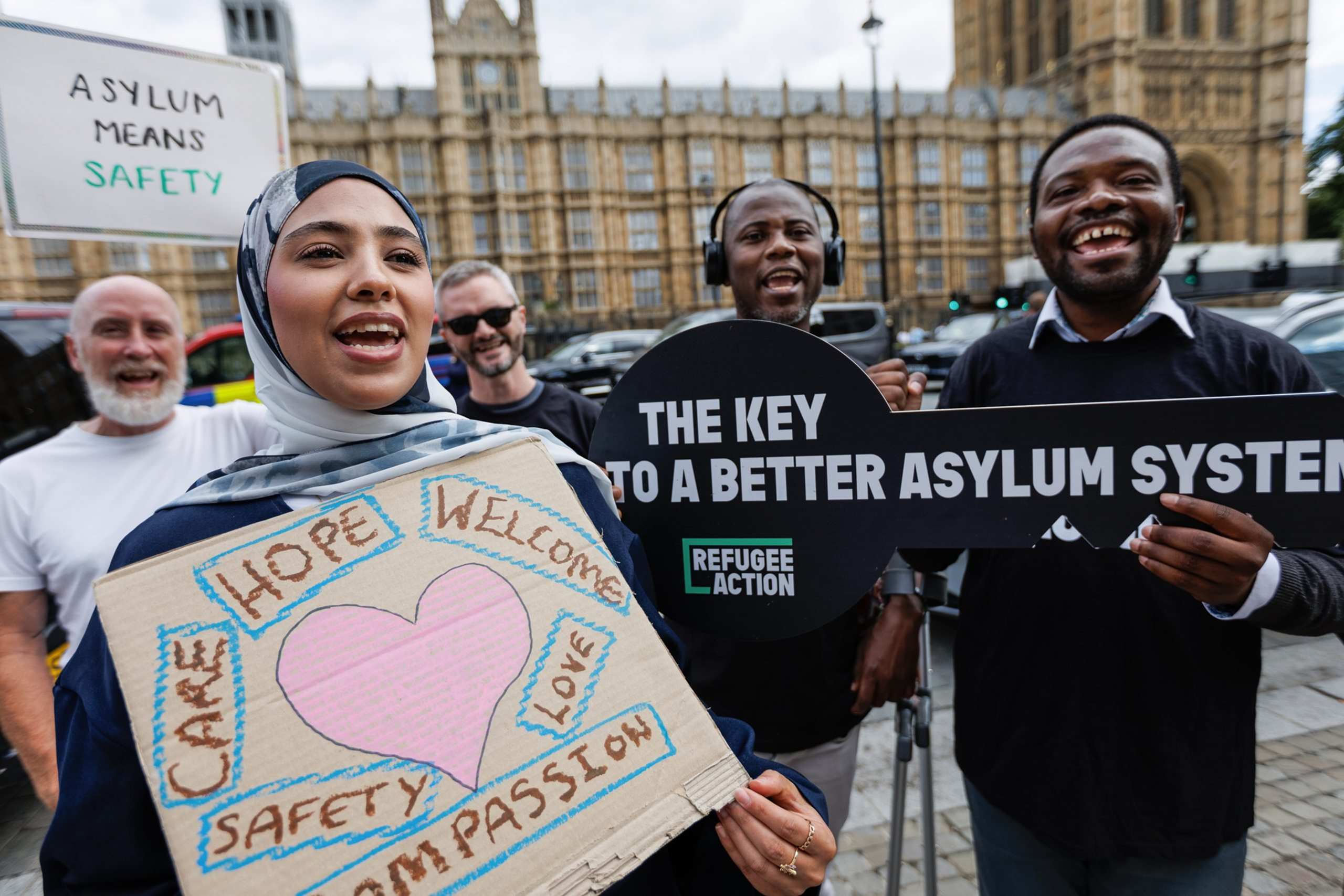 A group of campaigners are chanting down the camera lense. There's lots of energy in this photos. They hold signs which say "asylum means safety" and "The key to a better asylum system" which is written on a sign shaped like a key.